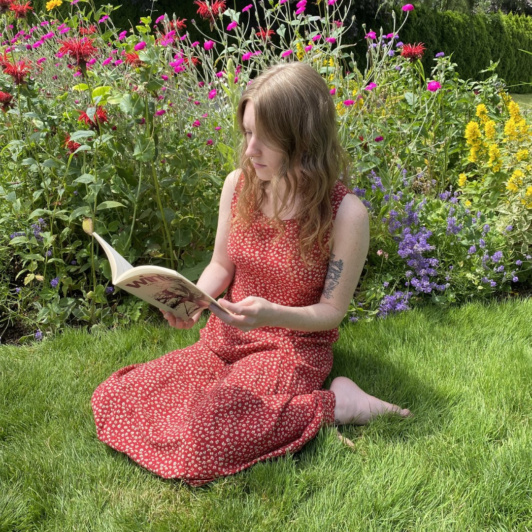 a woman sits in front of her flower garden, reading a book.